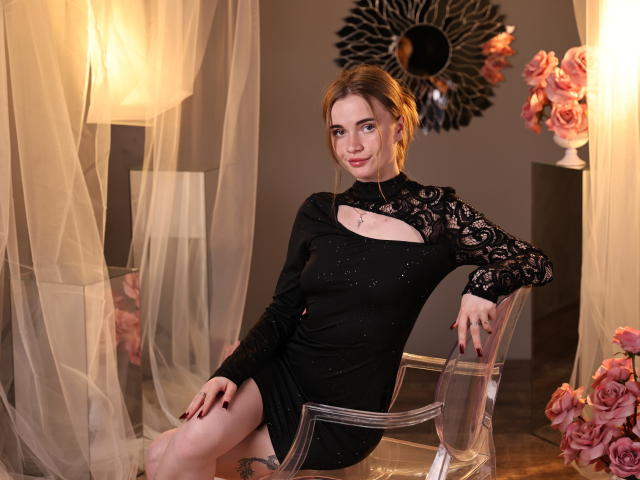 MargaritaKis, seated on a transparent chair, wearing a black lace dress with a cutout, legs apart, hand on chair. Warm lighting, pink roses, black sunburst mirror. Fucking sexy.