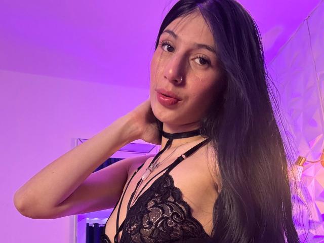 Photograph of SophieBeaumont, Asian woman with long black hair, wearing a black lace bra, looking over her shoulder with hand on neck. Purple and pink lighting in background, seductive expression.