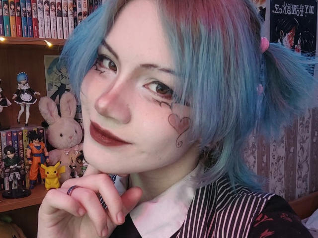 Photo en gros plan de KatlynRenteria avec des cheveux bleus attachés en couettes, un rouge à lèvres foncé, et des tatouages en forme de cœur sur les joues. Chemise rayée noir et blanc, main près de la bouche, figurines et peluches d'anime en arrière-plan. Tellement mignon..