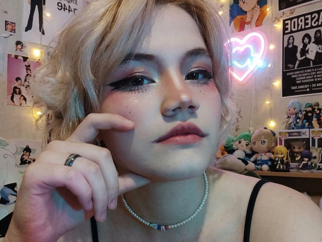 Close-up de KatlynRenteria com cabelo loiro, delineador marcante e um colar de pérolas, com o queixo apoiado na mão. Luzes de neon e pôsteres de anime ao fundo. Uma aparência incrivelmente fofa, ligeiramente sedutora..