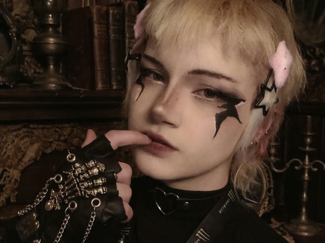 Close-up foto van KatlynRenteria met bleke huid, blond haar en zwarte ster-gezichtsschildering. Ze draagt een zwarte leren outfit, handschoenen zonder vingers met kettingen, en ze bijt verleidelijk op haar vinger..