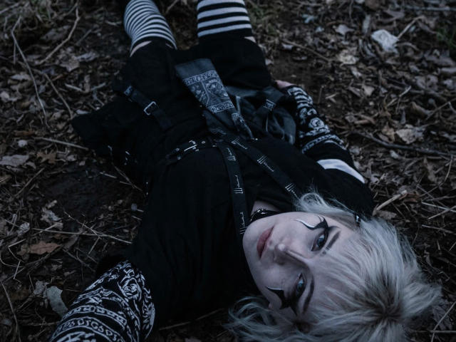 Photographie de KatlynRenteria allongée sur un sol sale, vêtue de vêtements à motifs noir et blanc, de chaussettes rayées et de chaussures noires. gants. Peau pâle, yeux bleus, cheveux courts argentés, regard intense, tension sexuelle.