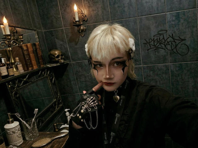 KatlynRenteria, avec sa peau pâle et ses cheveux blonds platine, son maquillage noir et sa veste en cuir noir, tenant un objet en argent. Elle porta son doigt contre ses lèvres. Pièce faiblement éclairée avec des bougies et des graffitis.