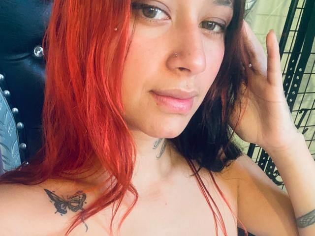 Nahaufnahme-Selfie von RoxanaSanchezz mit roten Haaren, heller Haut und einem Schmetterlingstattoo auf ihrer linken Schulter. Sie schaut in die Kamera, eine Hand im Haar, und trägt einen schwarzen Lederstuhl. Verdammt heiß..