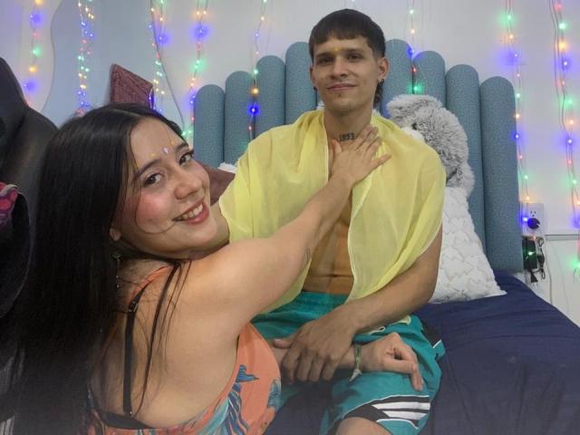 AnjaliRohan, con el pelo largo y negro, sonriendo, vestida con una blusa naranja con estampado floral, tocando a un hombre sin camisa con el pelo corto y castaño. pantalones verdes, camisa amarilla y un cabecero gris con luces de colores..