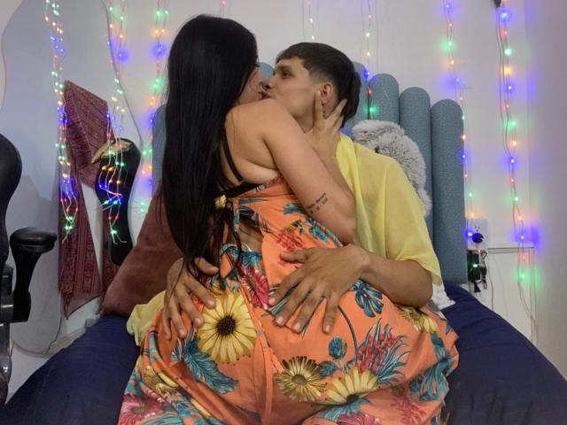 AnjaliRohan, con i suoi lunghi capelli neri, è seduta in grembo a un uomo e lo bacia appassionatamente. Indossa un abito floreale arancione vivace, che lascia scoperta la schiena. Indossa una camicia gialla e le afferra i glutei. Sullo sfondo, luci colorate..