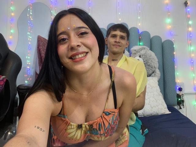 AnjaliRohan, com cabelo preto e pele morena, sorrindo, vestindo um top de biquíni colorido, inclinada para frente, expondo o colo. com um pênis circuncidado, semi-ereção, visível atrás dela, luzes coloridas em forma de cordão ao fundo..