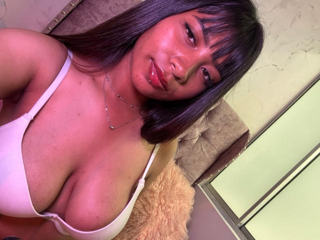 Close-up photo of NahomyNoir, a dark-skinned woman with long black hair and bangs, wearing a low-cut white tank top, revealing large breasts. She has a slight smile and glossy lips. Bright lighting.