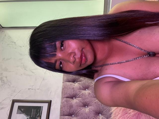Close-up of NahomyNoir, a young woman with long black hair and bangs, wearing a white tank top, showing a slight smirk. She's holding a camera, with a tufted bed behind her.
