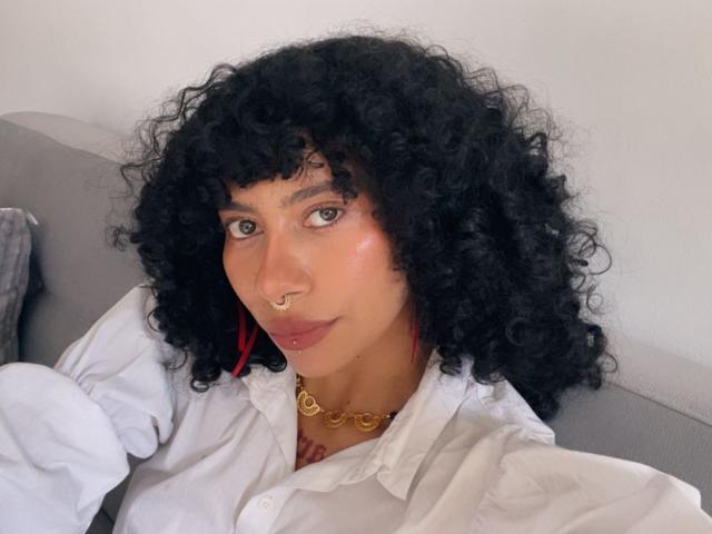 Fotografia de CharlotteXMonroe, uma mulher curvilínea com pele marrom média e cabelo preto encaracolado e curto. Ela está vestindo uma camisa branca, um colar de ouro e um piercing no nariz. Um olhar ligeiramente sedutor e direto..