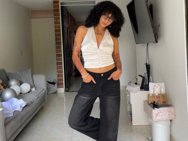 Fotografía de CharlotteXMonroe con cabello negro rizado, vistiendo un top blanco de tirantes y pantalones negros, con las manos en los bolsillos. De pie en una sala de estar con un televisor, un sofá y globos. Increíblemente atractivo/a..