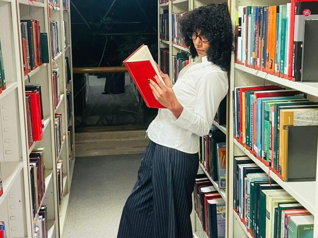 CharlotteXMonroe, cabello negro rizado, camisa blanca de manga larga, falda negra a rayas, sosteniendo un libro rojo, de pie entre estanterías, lomos de libros de colores, biblioteca, mirada intensa, sexy, concentrado, seductor, pasillo estrecho, iluminación brillante..