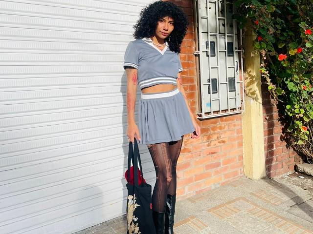 Photographie de CharlotteXMonroe debout devant un mur de briques, portant un haut court gris et une jupe, ainsi que des bas résille noirs. des bas, tenant un sac noir. Cheveux bouclés, pose confiante, tension sexuelle.