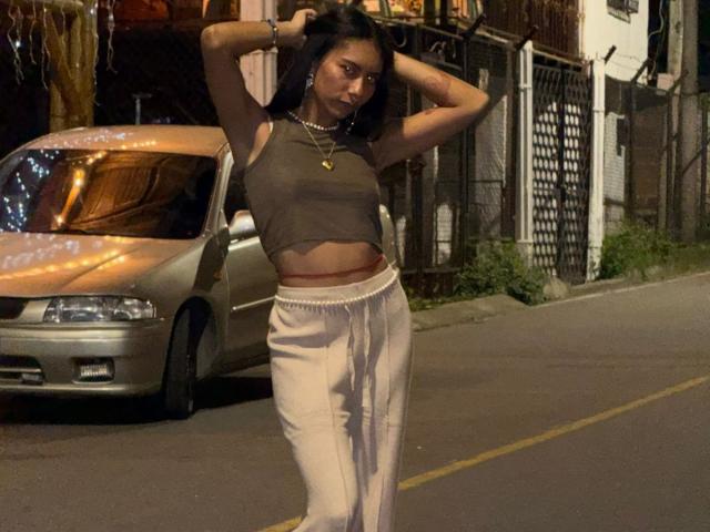 Foto noturna de CharlotteXMonroe, em pé em uma rua, com as mãos atrás da cabeça, vestindo uma blusa marrom curta, expondo parte superior do corpo, calças brancas, colar, carro com luzes laranja ao fundo. Tensão sexual.