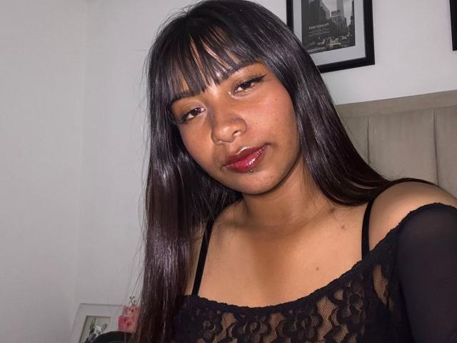 Gros plan de NahomyNoir, une femme latino-américaine aux longs cheveux noirs et à la frange, portant un haut en dentelle noire, du rouge à lèvres et expression sensuelle.
décolleté visible, tension sensuelle, regard séduisant.