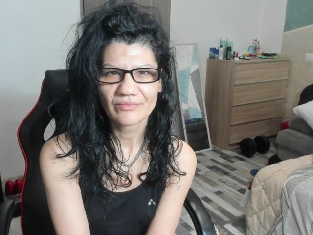 Vanessa69Xo, con i capelli neri spettinati e gli occhiali, indossa una canotta nera, è seduta su una sedia nera e guarda la fotocamera. con un sorriso appena accennato. Camera da letto, comò disordinato, letto..