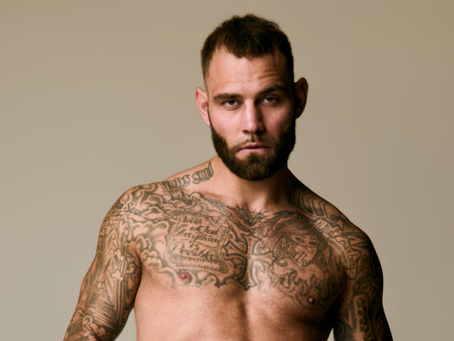 Fotografia di AlphamanJay senza camicia, con una corporatura muscolosa, numerosi tatuaggi che coprono il petto e le braccia, capelli corti e scuri. capelli e barba. Fissa intensamente in avanti, emanando una forte tensione sessuale..