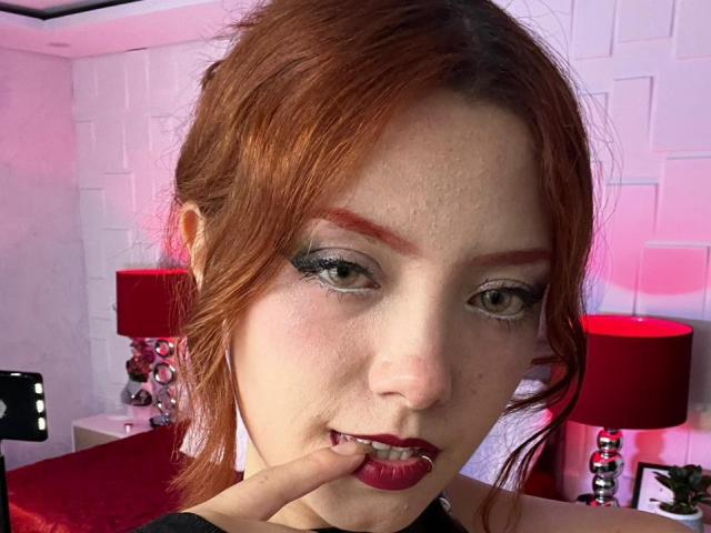 Close-up foto van Emma Thomson met rood haar, smoky oogmake-up en rode lippenstift. Ze bijt in haar vinger, en kijkt verleidelijk met een halfglimlach. Op de achtergrond staan rode lampen en een witte bakstenen muur..
