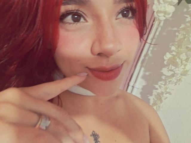 Close-up photo of MiaLeroy with red hair, fair skin, and brown eyes, biting her finger with a slight smile. Visible tattoo on her chest. Hand with ring on right. Fucking sexy.
