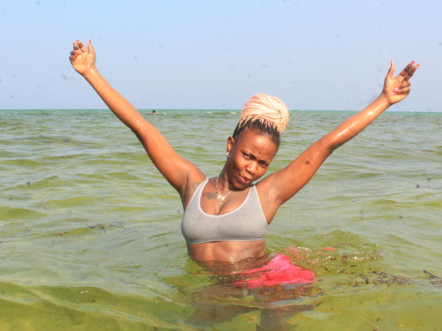 StylishAurora, une femme à la peau foncée avec des cheveux blonds tressés et mouillés, se tient dans une eau de mer verte, les bras levés. Elle porte un soutien-gorge de sport blanc, qui brille d'eau. Un maillot de bain rouge est visible en dessous..