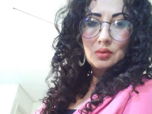 Close-up de Samantha Antolin, com cabelo preto encaracolado, usando óculos redondos e um casaco rosa. Ela tem uma expressão sedutora, com lábios vermelhos, olhando diretamente para a câmera, transmitindo uma forte tensão sexual..