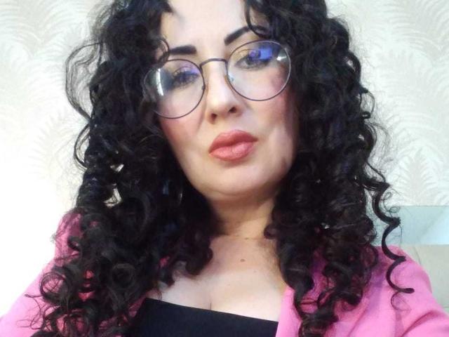 Nahaufnahme von Samantha Antolin mit lockigem, schwarzem Haar, die runde Brillen trägt und einen pinken Pullover über einem schwarzen Oberteil anhat. Sie hat volle Lippen, helle Haut und einen leicht provokanten Ausdruck..