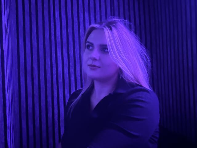 Fotografia de uma modelo PlusSizeBeauty com cabelos longos e azuis brilhantes, vestindo uma jaqueta preta, em frente a um fundo vertical azul escuro. Fundo listrado. A luz azul intensa realça seu rosto e cabelo. Expressão sensual..