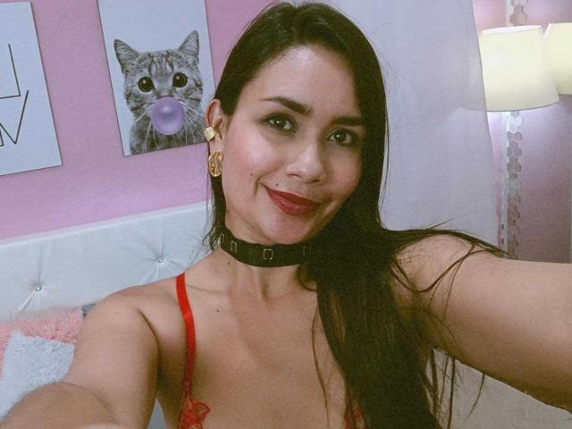 SophieJones69, com cabelo castanho escuro, roupa íntima vermelha, colar preto, batom vermelho, sorrindo para a câmera, sexy, em uma cama branca. Almofada rosa, pôster de gato com tema de chiclete, luminária ao fundo. Muito quente.