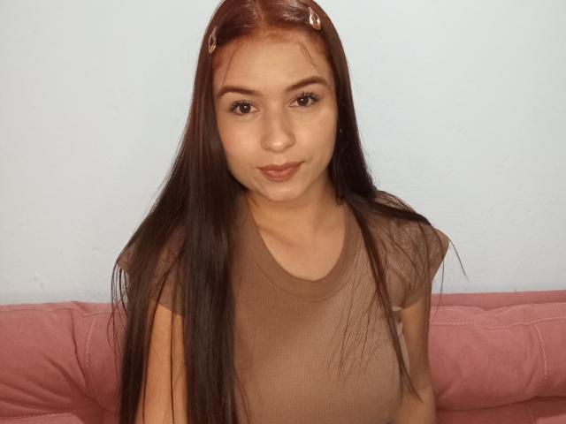 Fotografia de Lyra4, uma jovem com longos cabelos pretos, vestindo uma blusa marrom transparente, sentada em um sofá rosa. Ela tem olhos castanhos, pele clara e um sorriso ligeiramente sedutor..