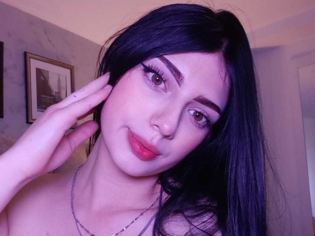 Close-up van Isabella Rossi, bleke huid, zwart haar, rode lipstick, dikke wenkbrauwen, lichte glimlach, hand die wang aanraakt, draag een zilveren ketting, kamer met gedimd licht, ingelijst beeld aan de muur, zacht paars licht. Heel aantrekkelijk..