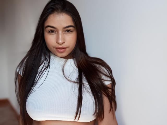 Photograph of Lyra4, a dark-haired woman with long, straight hair, wearing a white, ribbed, sleeveless top that highlights her large breasts. She has an intense, seductive gaze. Bright, even lighting.