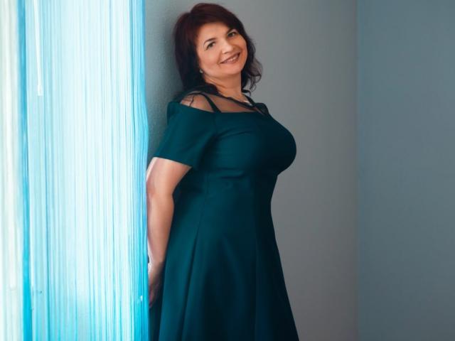 LedyNika, eine Frau mittleren Alters mit dunkelbraunen, mittellangen Haaren, steht vor einem hellblauen Hintergrund und trägt ein schwarzes kleid mit offener Schulter, wobei ihre großen Brüste deutlich sichtbar sind, lächelnd und selbstbewusst. Offensichtliche sexuelle Spannung..