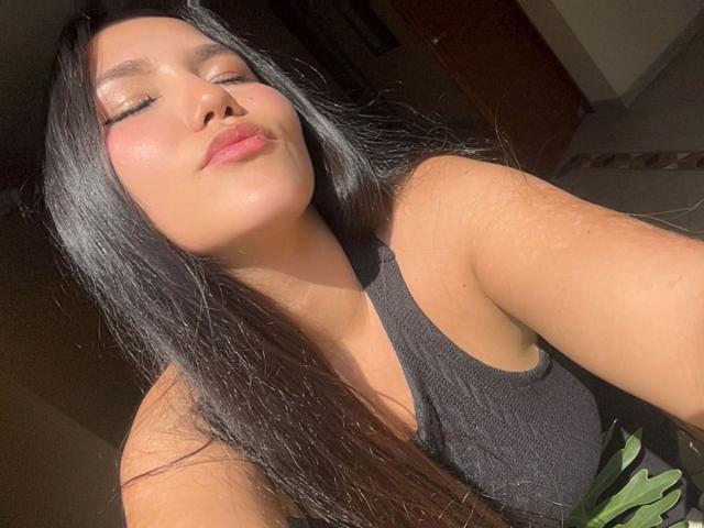 Primer plano de MariaRowe, una atractiva mujer asiática con cabello largo y negro, que lleva un top de tirantes negro, con el rostro iluminado por el sol, con los ojos cerrados. labios ligeramente fruncidos, luz solar intensa, fondo sombreado, pose informal.