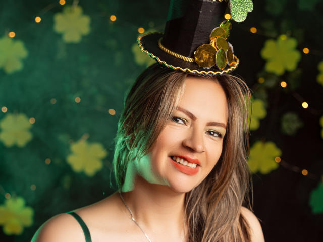 Close-up of LadyLure with a sexy smile, wearing a black top hat with gold trim and a small gold charm. Green and yellow flowers in the blurred background. Fucking hot.