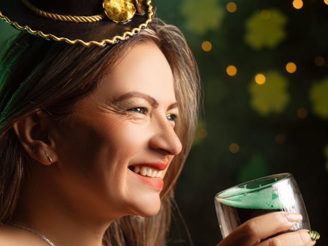 Close-up photo of LadyLure, smiling with red lips, holding a glass of green liquid. She wears a gold headpiece with a heart-shaped jewel, dark hair, dim lighting, bokeh background.