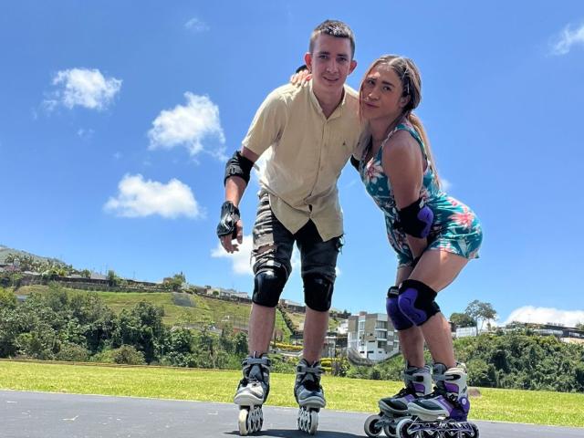 MyaAndJack69, um homem magro com cabelo castanho curto, vestindo uma camisa bege e shorts pretos, e uma mulher morena curvilínea vestida com roupas coloridas. vestido, em patins, inclinada para frente, sorrindo. Céu azul brilhante.