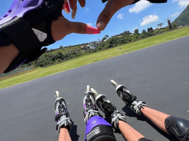 Close-up photo of MyaAndJack69's legs in black and white running shoes, wearing purple shorts and black knee pads, on an asphalt road with grass and trees in the background. A hand holds a red pen above.