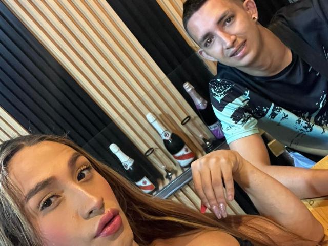 Photographie de deux personnes, MyaAndJack69 avec de longs cheveux bruns et des lèvres pulpeuses au premier plan, et une autre MyaAndJack69 avec des cheveux courts et foncés et un t-shirt noir, une personne penchée en arrière-plan. Tension sexuelle.