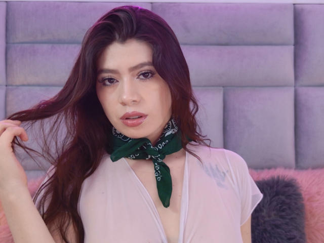 Fotografia de Linda Mancini com cabelos longos e castanho-escuros, vestindo uma camisa branca com decote em V e um lenço verde na cabeça, segurando o cabelo. com uma expressão sedutora. O fundo inclui uma parede acolchoada cinza..