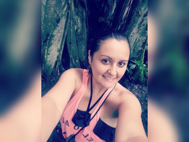 Photograph of LaurenSpitz, wet, smiling, taking a selfie, wearing a pink and black tank top, dark hair pulled back, against a textured, blurred tree background, suggestive, athletic, wet skin.