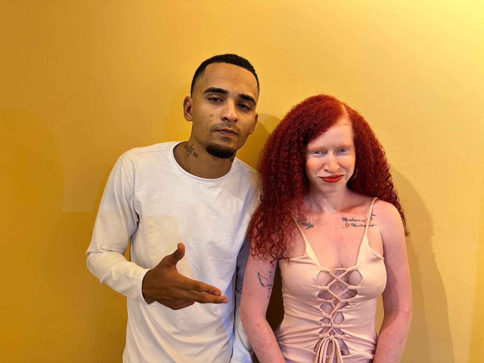 Photographie d'un duo noir et blanc. Un homme noir aux cheveux courts, portant une chemise blanche à manches longues, faisant un geste de la main droite. Femme blanche aux cheveux roux, tatouages sur les bras, haut en corset rose, appuyée contre un mur jaune..