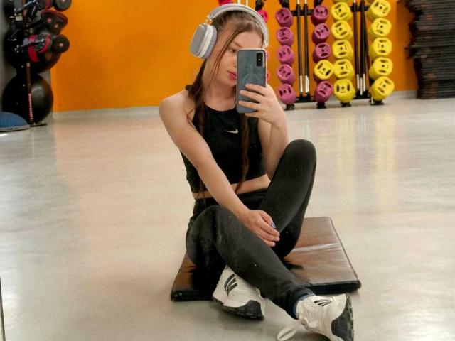 VioletaNova, una mujer delgada con cabello largo y castaño, está sentada en un banco en un gimnasio, vestida con ropa negra y zapatillas blancas. Tomando una selfie con un teléfono inteligente, con auriculares puestos. Tensión sexual evidente..