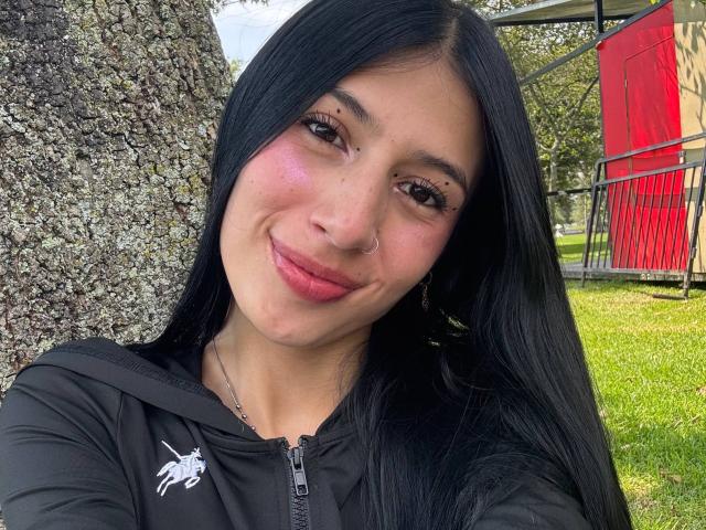 Photograph of SophieBeaumont with long black hair, light skin, and a nose ring, smiling at the camera. She wears a black hoodie with a white graphic, in an outdoor park. Red play structure in the background.