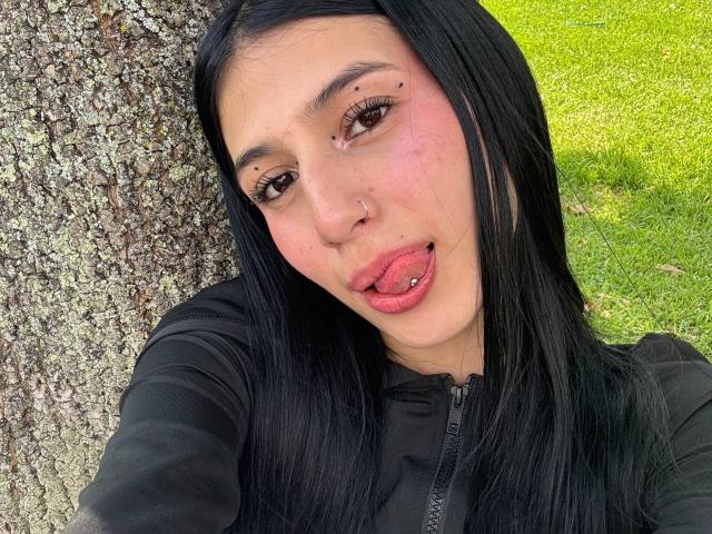 Gros plan de Sophie Beaumont, une femme latina aux longs cheveux noirs, qui tire la langue et mord sa lèvre inférieure. Elle porte un sweat à capuche noir avec fermeture éclair, et on peut voir une petite tâche de rousseur au-dessus de sa lèvre. L'arrière-plan est un gazon jaune vif..