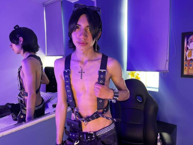 Aetternum with dark, shaggy hair, wearing black leather harness and belt, exposed chest with cross necklace, sitting in front of mirror, blue and purple lighting, looking seductive, cock strap visible.