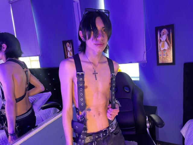 Aetternum staat voor een spiegel, zonder shirt, met zwarte leren ophangers en een riem, en er is blauw licht. Spiegel reflecteert de rug. Tattoo op de borst. Posters aan de muur. Heel erg aantrekkelijk..