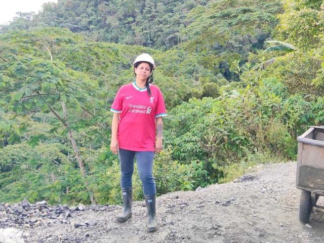 IsabellaSweett stands on a gravel road wearing a red shirt, blue jeans, black boots, and a white cap. Lush green forest in the background, with a rusty truck on the right. Fucking hot.