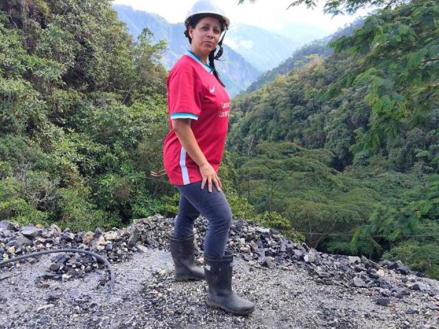 IsabellaSweett stands on rocky terrain, wearing a red shirt, black pants, and boots. She's in a forested area with lush green hills in the background. Her pose is confident and sexy.