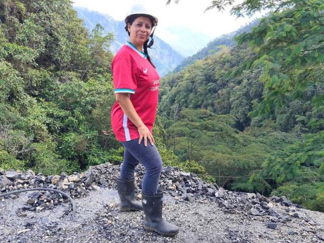 IsabellaSweett está de pie en un camino rocoso, vistiendo una camisa roja, pantalones negros y botas. Ella mira por encima del hombro con una ligera sonrisa. Un denso bosque verde en el fondo..