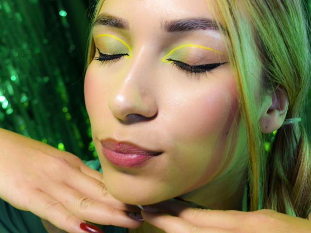 Primer plano de TanniaMoon con los ojos cerrados, sombra de ojos amarilla y negra, y lápiz labial rojo. Ella está tocándose la barbilla con dedos cuidadosamente arreglados, y las luces verdes se reflejan en su rostro. Absolutamente precioso/a..